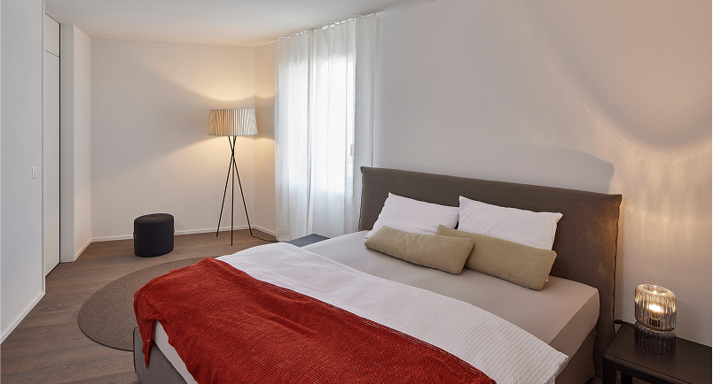 Ruhiges Schlafzimmer mit grossem Fenster, textilen Akzenten und warmem Licht.